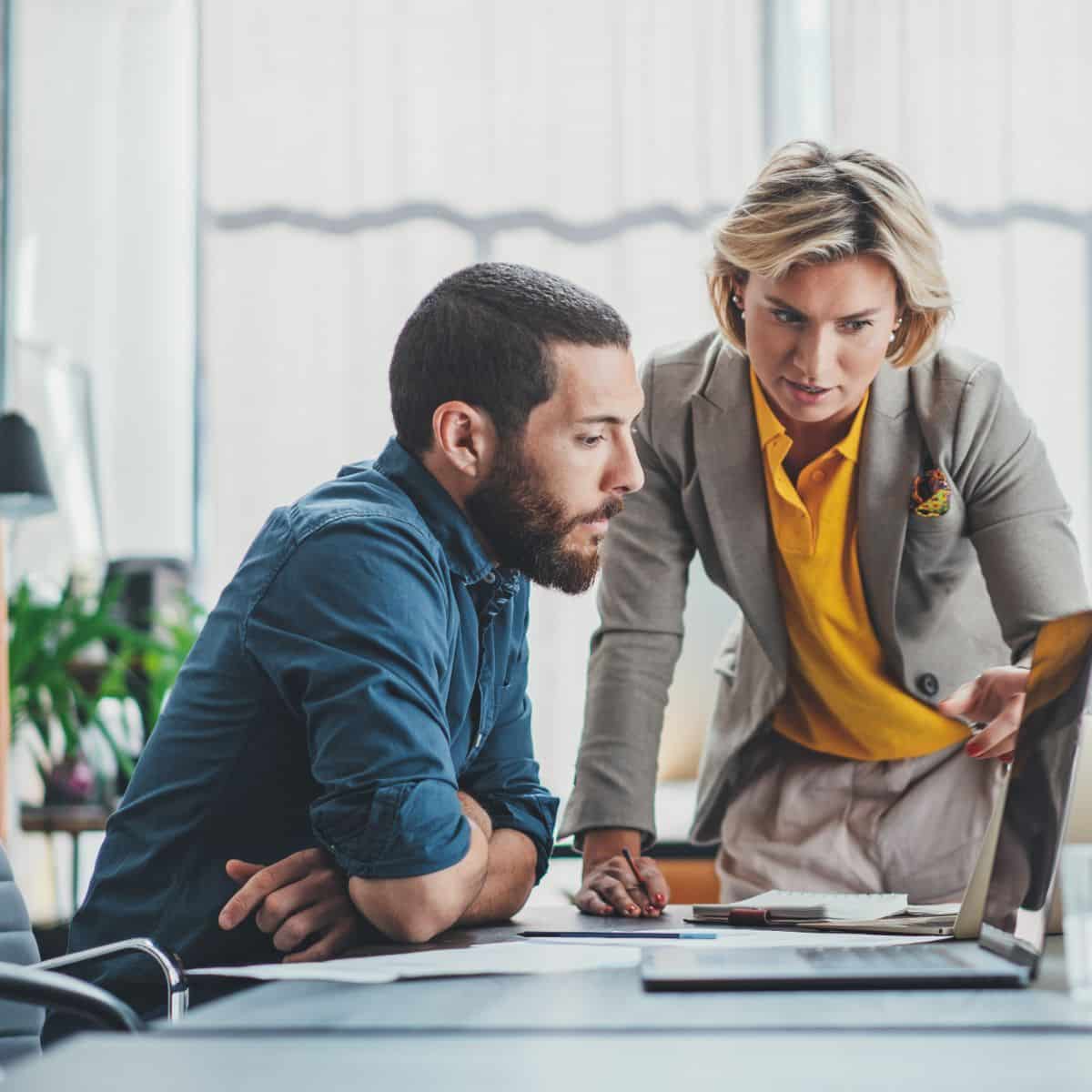 A man and woman in an office looking at a laptop screen.